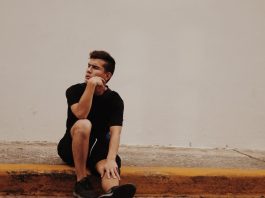 photo of man with hand on chin sitting on concrete pavement in front of white wall